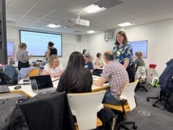 Classroom full of people sitting at desks and standing, discussing and working together.
